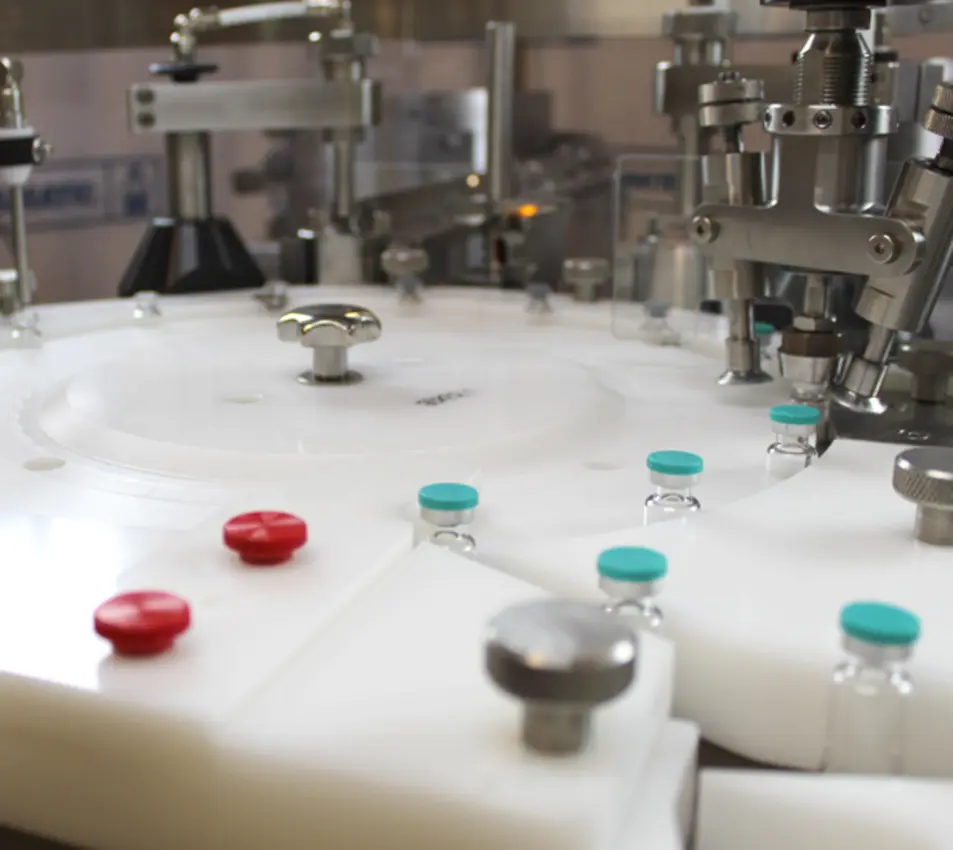 Close-up of a beverage filling machine with white carousel, metal nozzles, and red and teal capped containers.