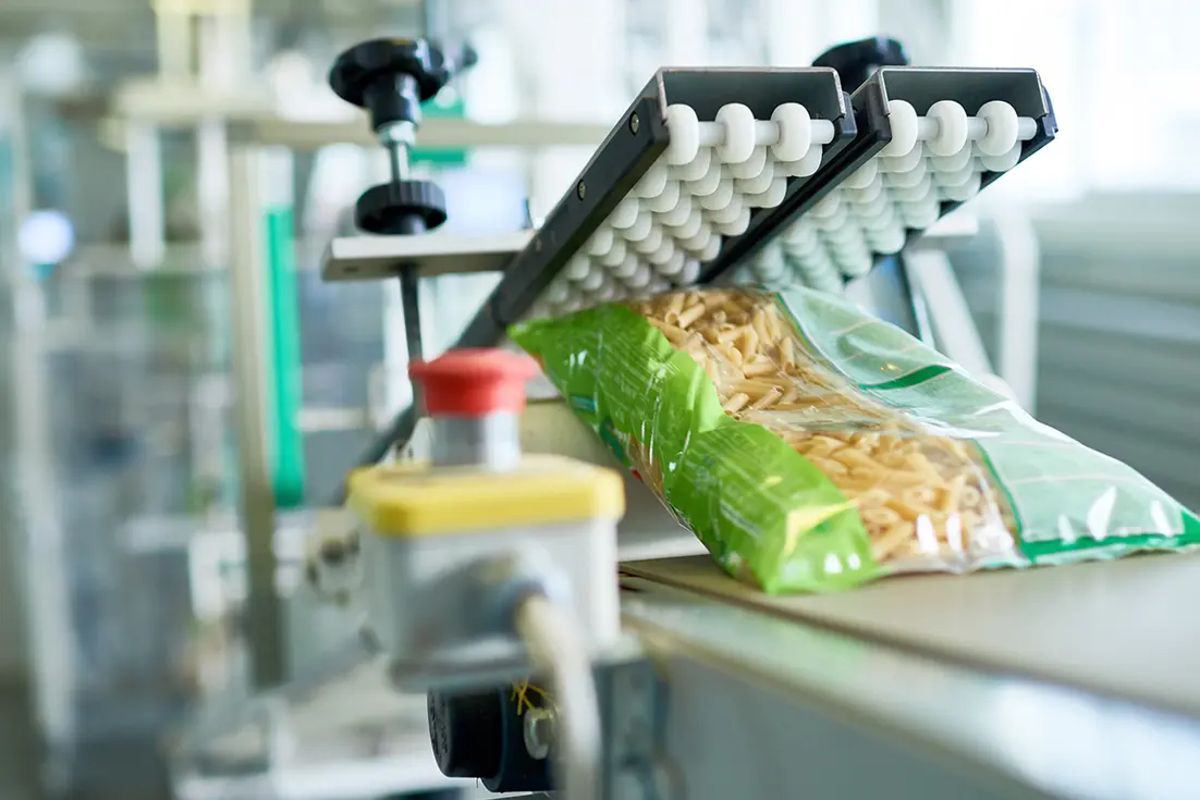 Green cook-in pouch of pasta on a conveyor beneath an industrial sealing or packaging machine.