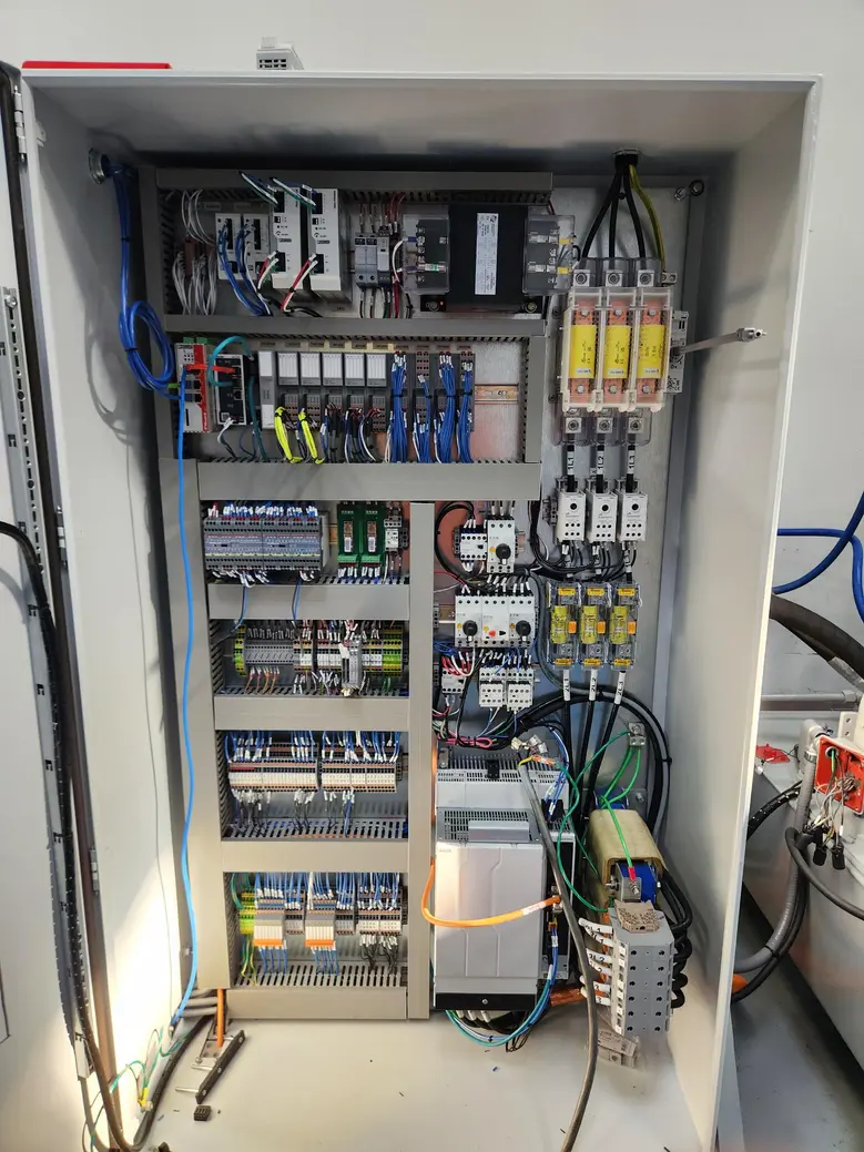 Open electrical control cabinet containing PLC modules, terminal blocks, wiring, circuit breakers, and cooling components.