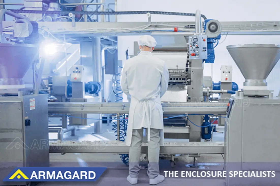 Worker in protective clothing stands among stainless steel processing machinery, conveyors, pipes, and electrical enclosures in a food plant.