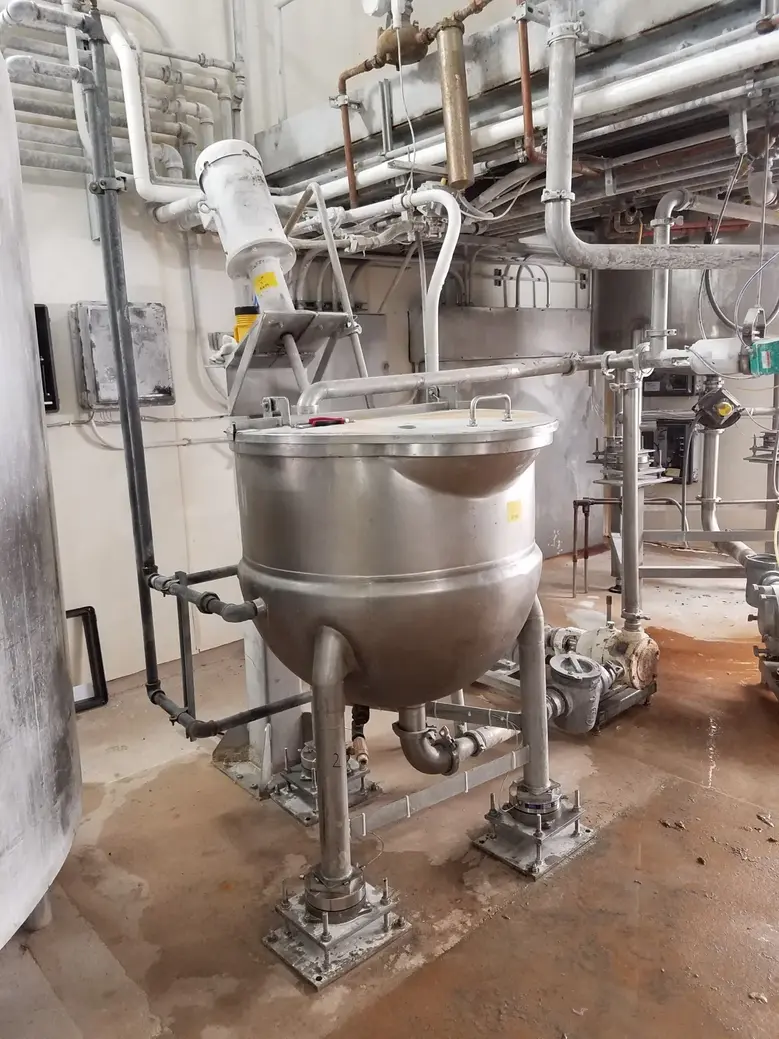 Stainless steel processing vessel with lid, valves, pipes, pumps, and surrounding industrial food-processing equipment.