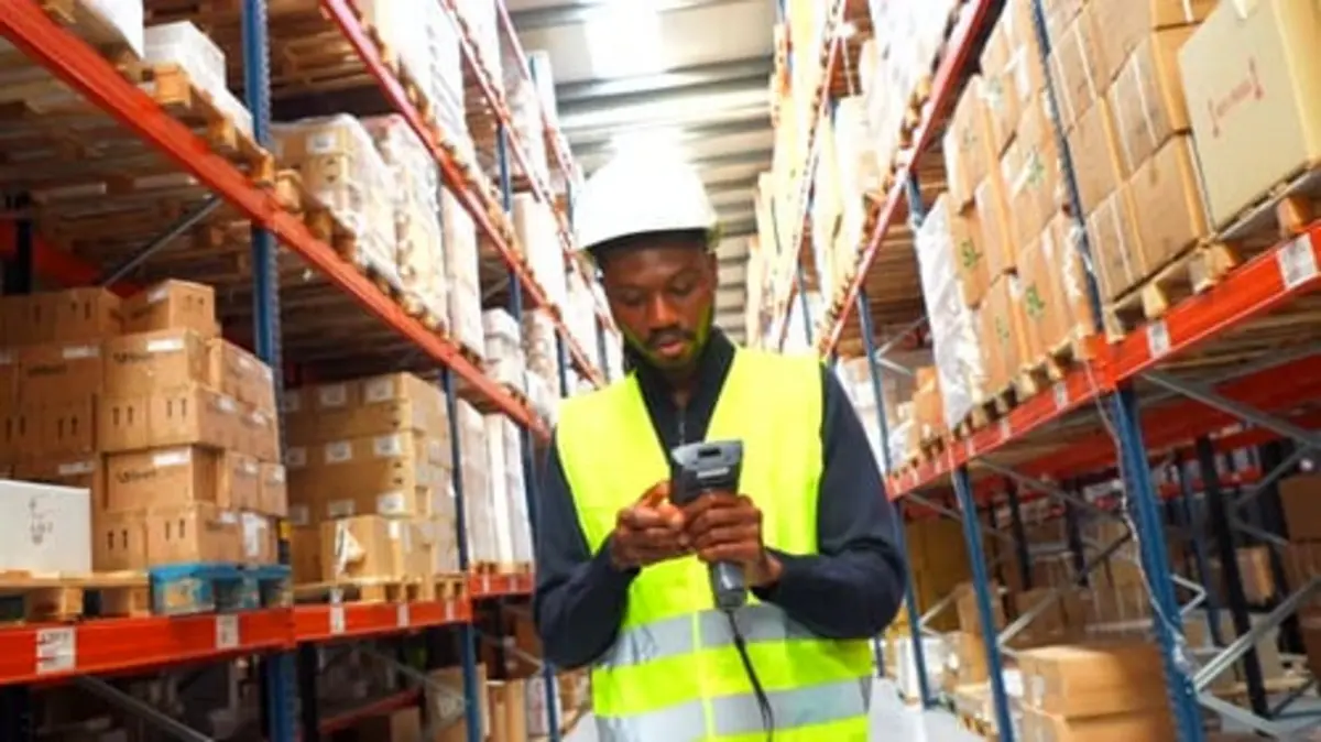 Warehouse worker in a hard hat and reflective vest scanning a barcode among shelves of boxes.