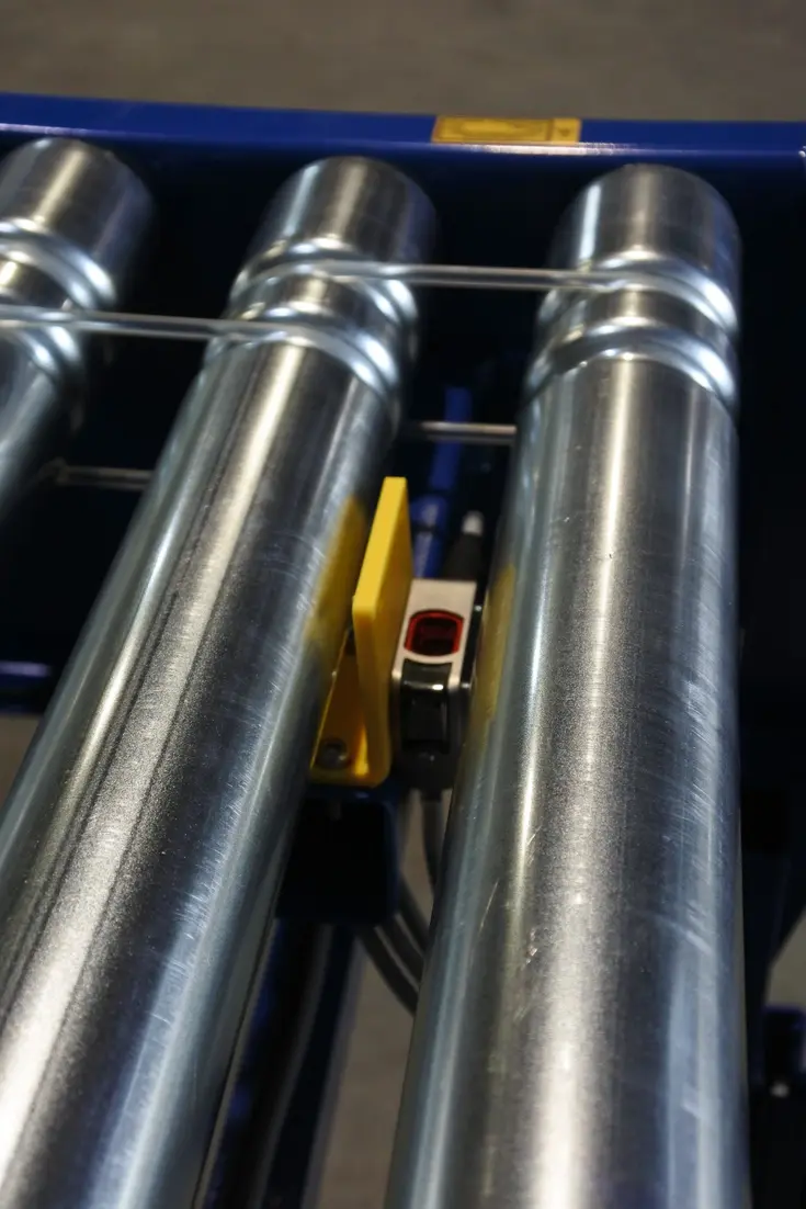 Close-up of stainless steel rollers with a small optical sensor and yellow guard positioned between them.