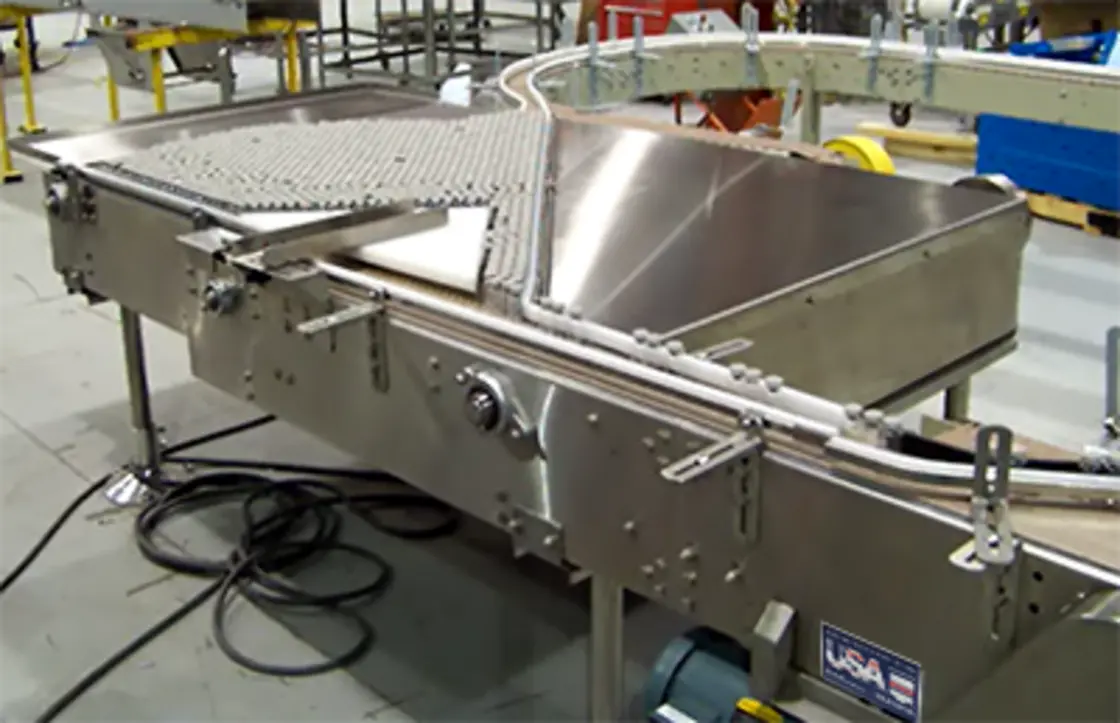 Stainless steel conveyor system with a curved accumulation table, guide rails, and industrial equipment in the background.