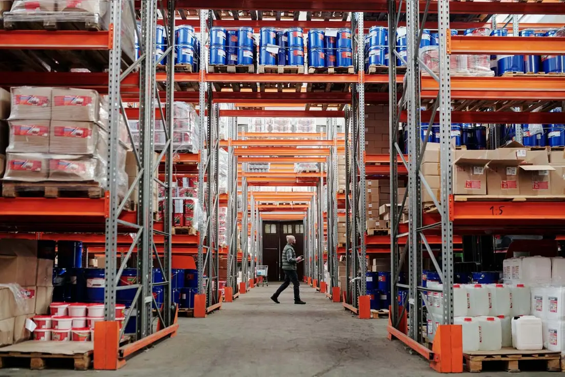 Warehouse aisle with stacked containers and boxes, workers, and palletized goods arranged on tall industrial shelving.
