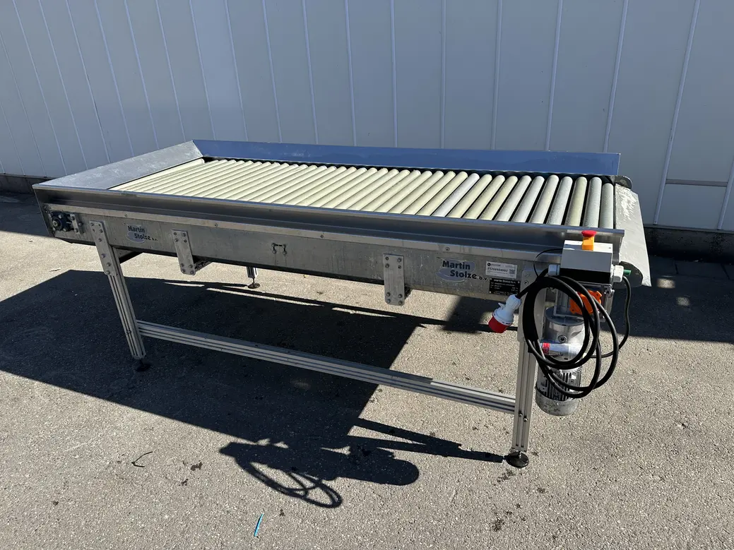Stainless steel roller conveyor with adjustable legs and hanging electrical cables, photographed outdoors beside an industrial building.