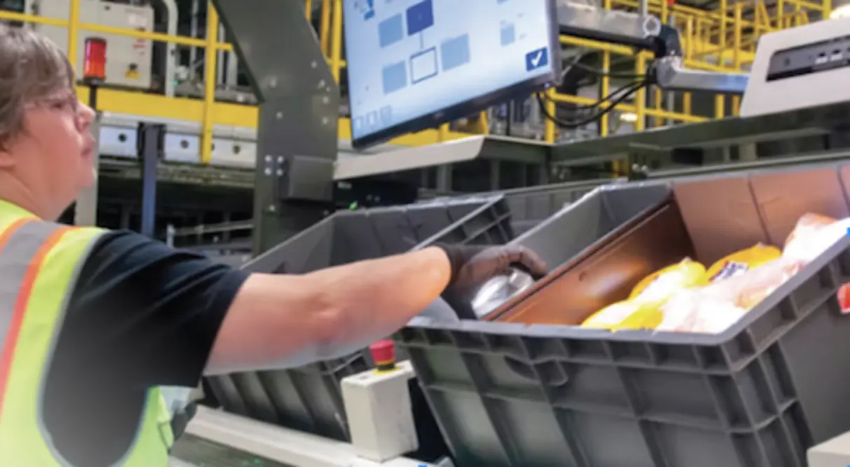 Worker inspects containers on a conveyor line beside a touchscreen control panel.