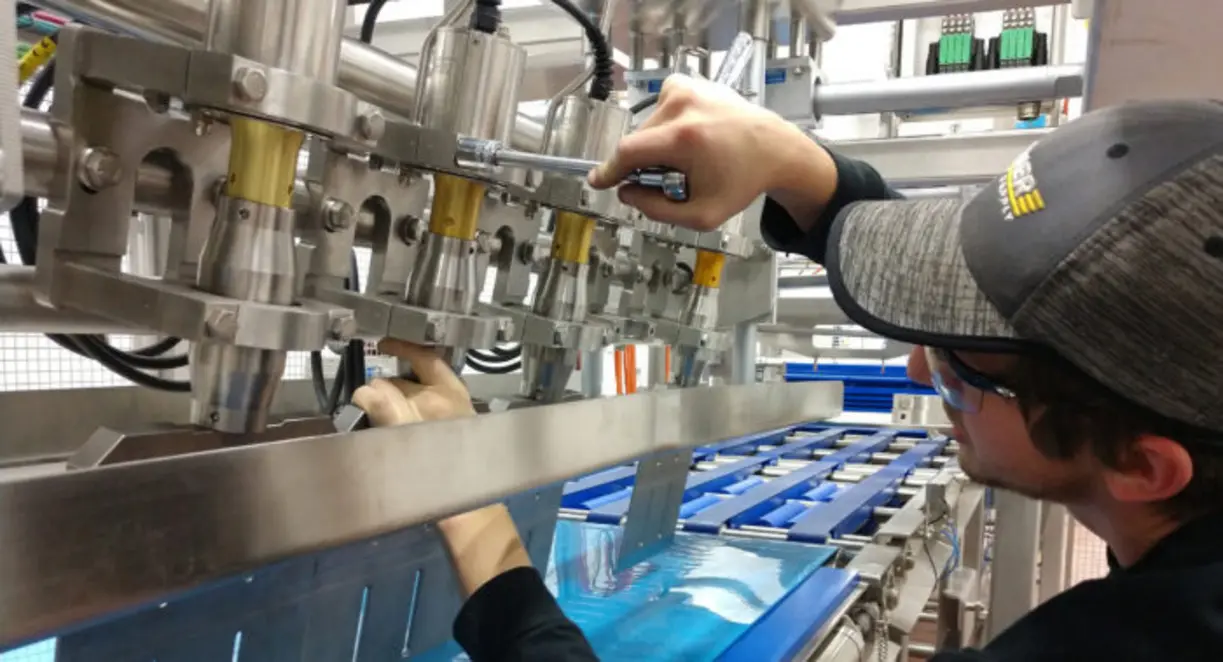 Technician adjusts a stainless steel food processing machine above a conveyor carrying blue trays.